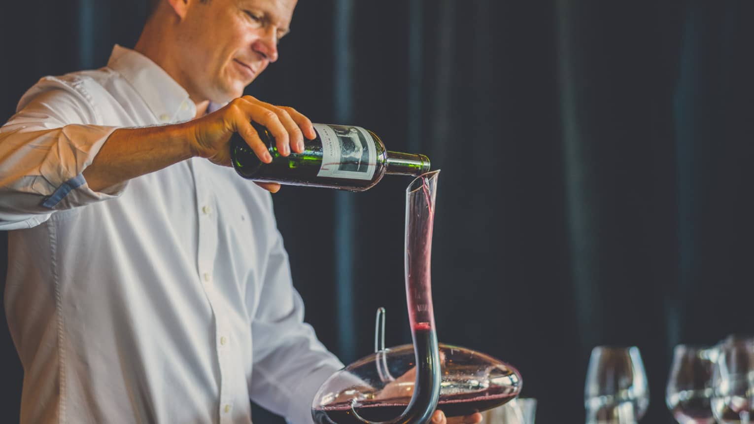 Man pours red wine into long, curved glass carafe near wine glasses