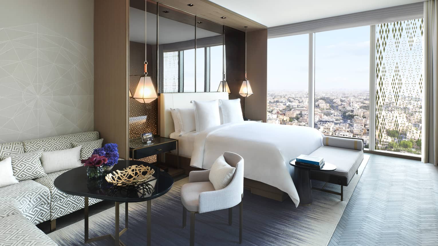 Hotel room L-shaped corner sofa, coffee table and chairs beside bed, wall of windows