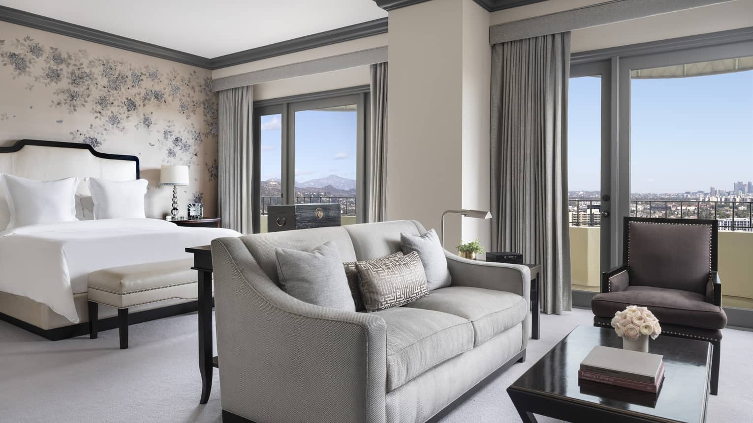 A hotel room with a coffee table, love seat, large bed with white sheets and two large windows.
