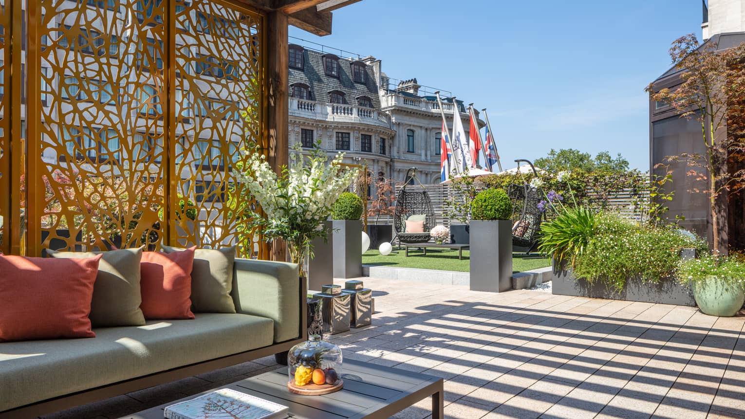 The Terrace Suite at Four Seasons London Park Lane in Mayfair features an expansive and beautifully designed outdoor terrace. The space is adorned with modern outdoor furniture including a sleek sofa with plush cushions and a low coffee table, perfect for relaxation or entertaining. The terrace is surrounded by lush potted plants, sculptural elements, and features decking that adds to the sophisticated ambiance. The view includes the tops of surrounding historic London buildings, creating a luxurious and serene urban garden atmosphere.