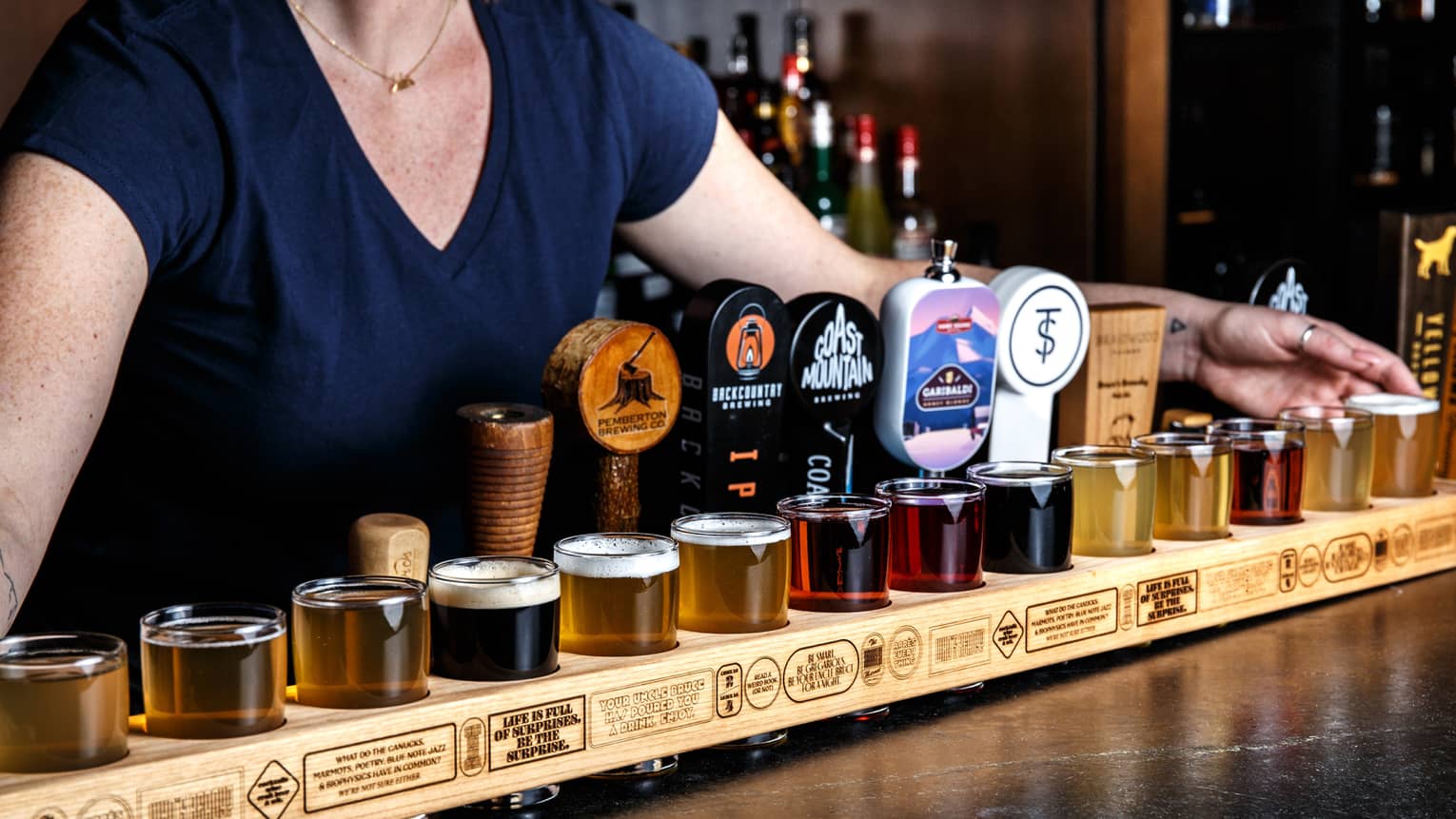 A series of glasses sit on a black table, with a female bartender standing behind.