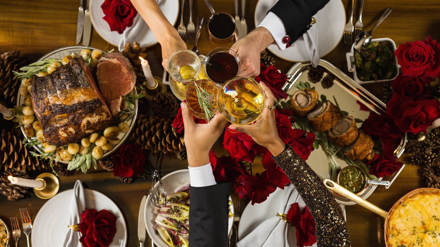 Four people hold their cocktail glasses together in a toast in the centre of a table filled with holiday dishes