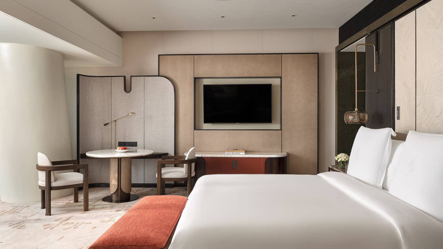 Contemporary hotel room with king bed, red cushioned bench, round table with two chairs and TV