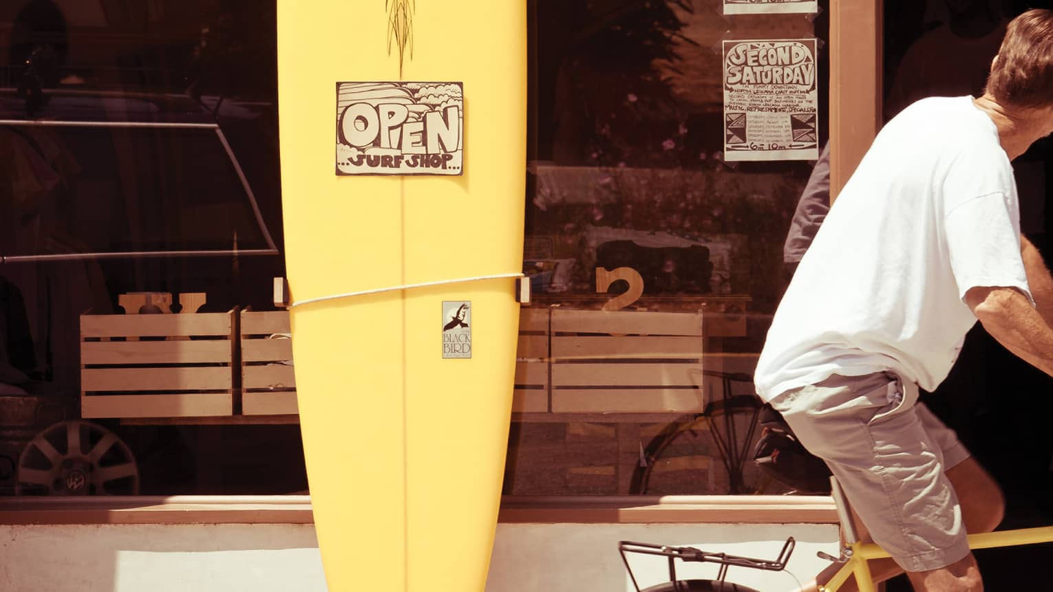 In the sun’s warm glow, a cyclist turns their head while riding by a yellow surfboard resting against a surf shop’s window. 