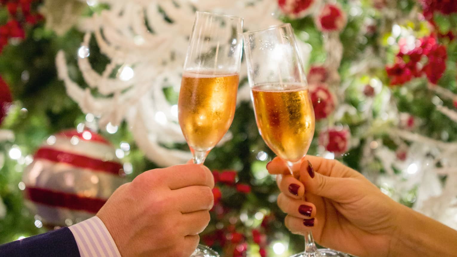Two hands toasting Champagne glasses with holiday decor in backdrop