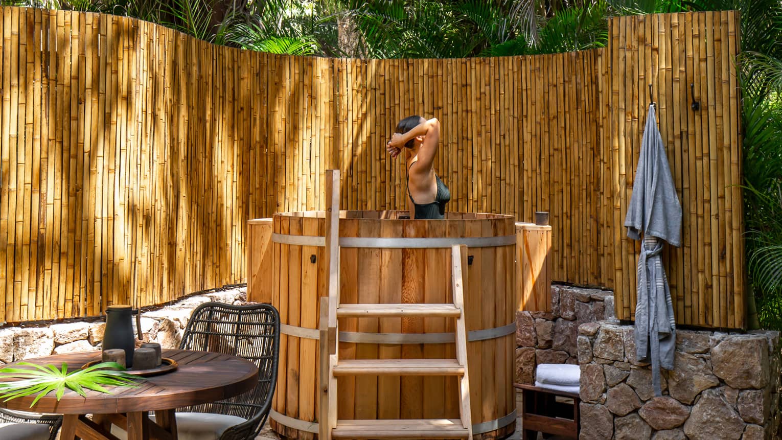 Woman emerges from soak in natural wooden outdoor bath, bamboo fence and palm trees surrounding