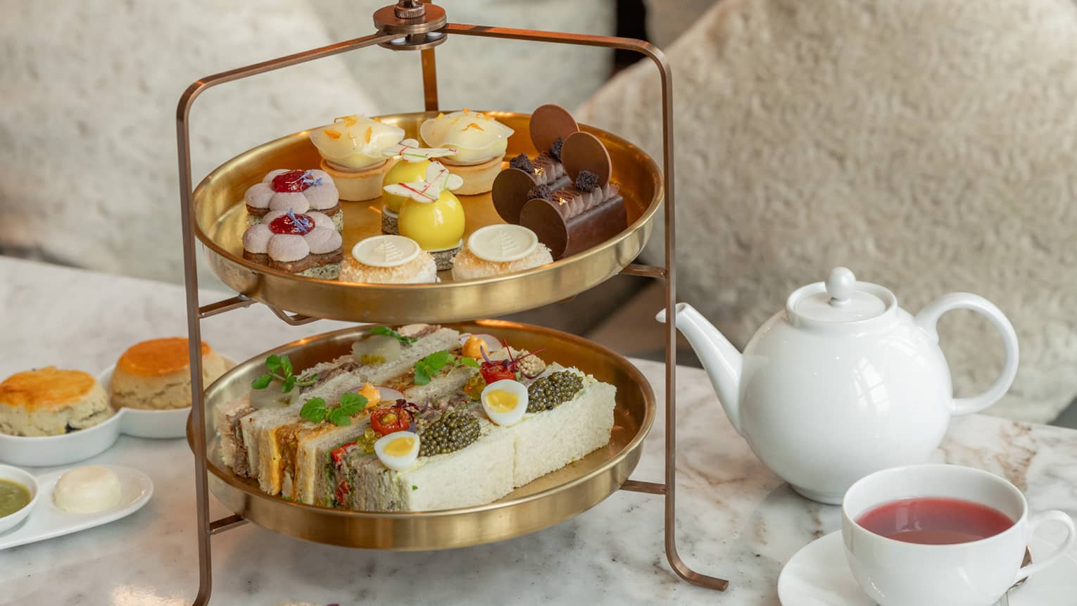 Two-tiered afternoon tea spread on marble coffee table in front of tan sofa