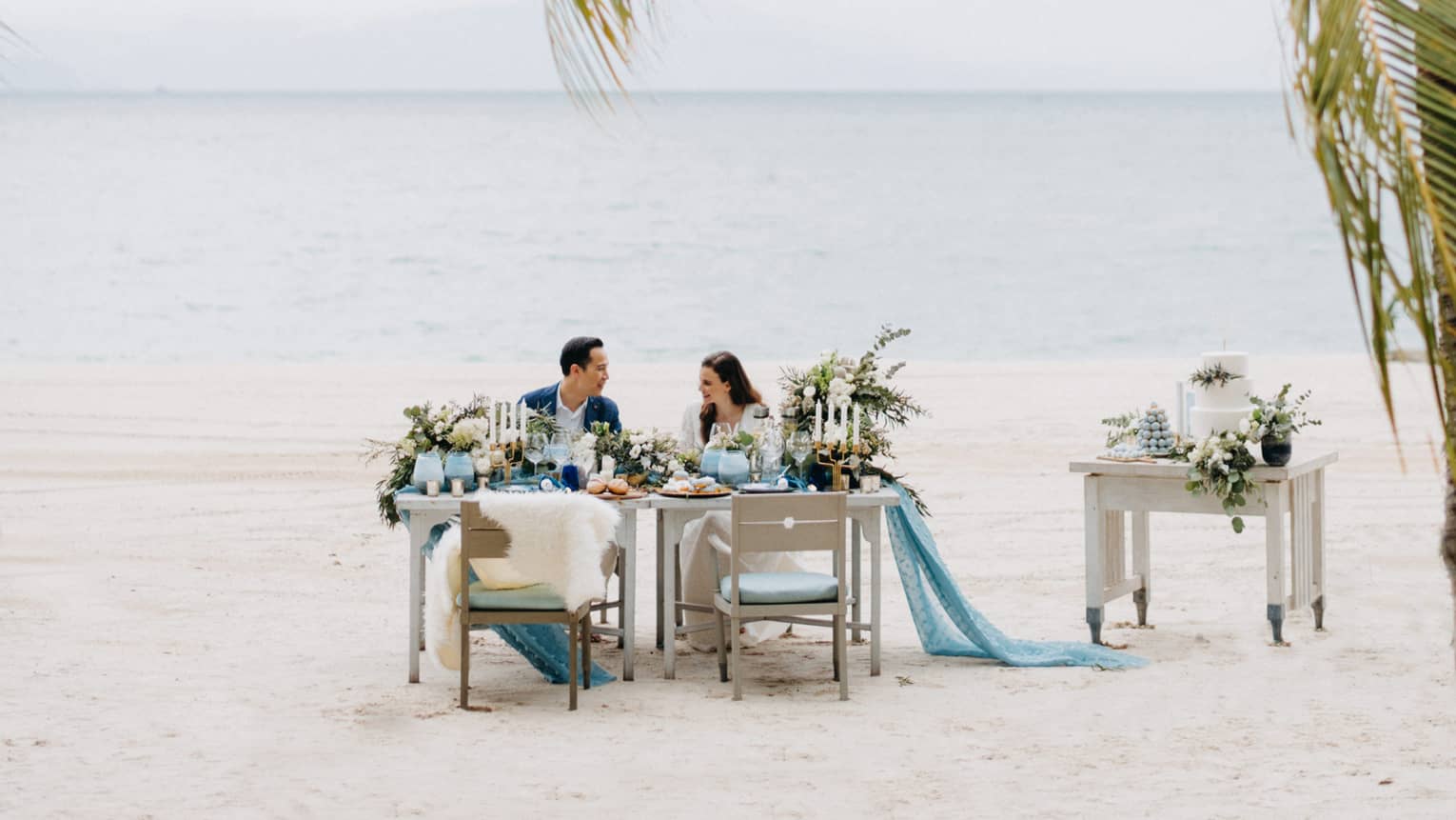 Smiling couple at private dining on white sand beach past palm leaves