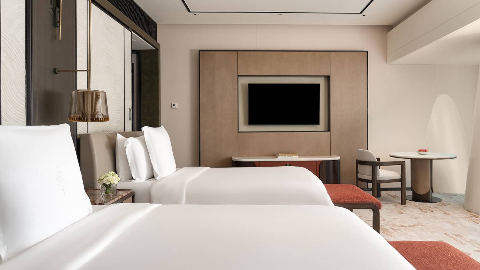 Hotel room with two beds, each with a red cushioned bench at the foot, plus a TV and a small table with chair