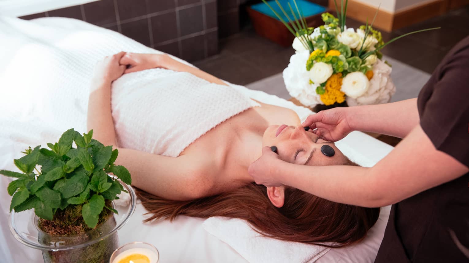 Spa staff places black stones on woman's forehead, cheeks as she lies under white sheet on massage bed