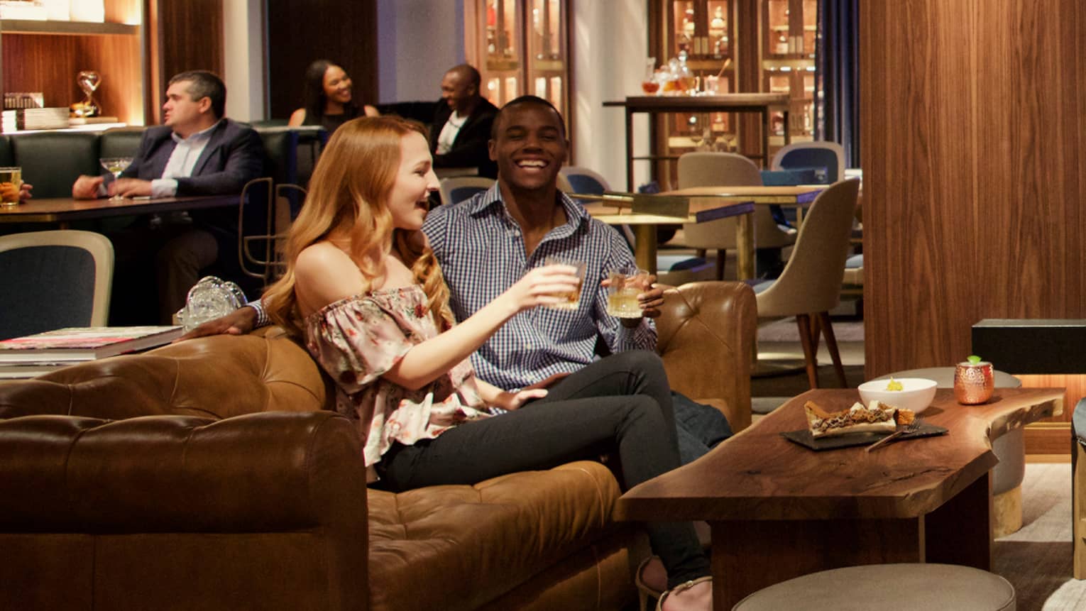 A man and woman sitting on a couch in a bar area drinking and laughing.