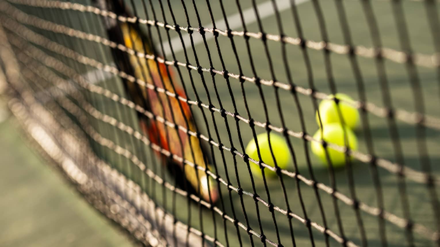 Close-up of tennis racket against net, three green tennis balls on ground