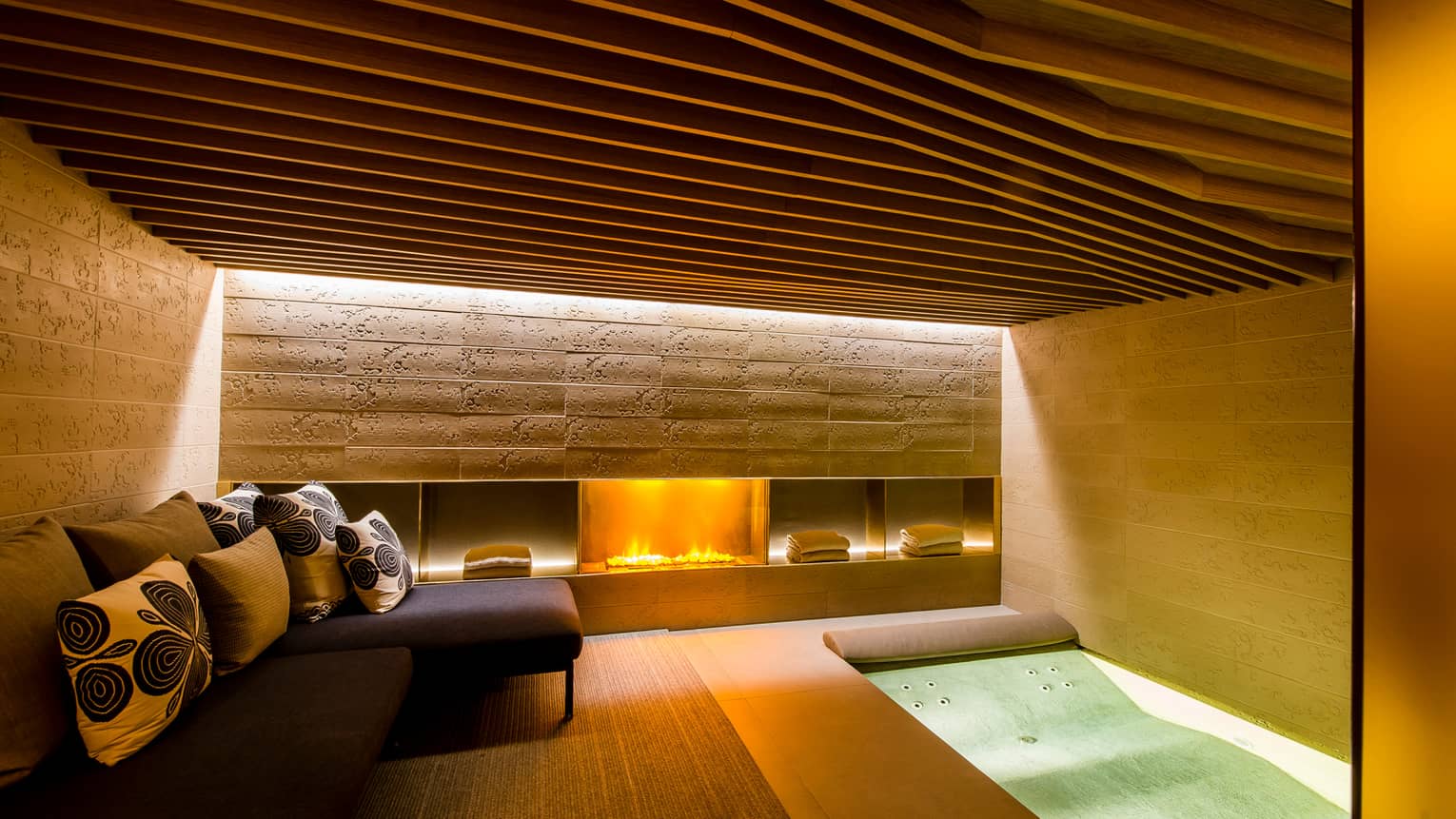 Gas fireplace in under stone wall in Urban Spa, small L-shaped sofa, cushions, spa tub