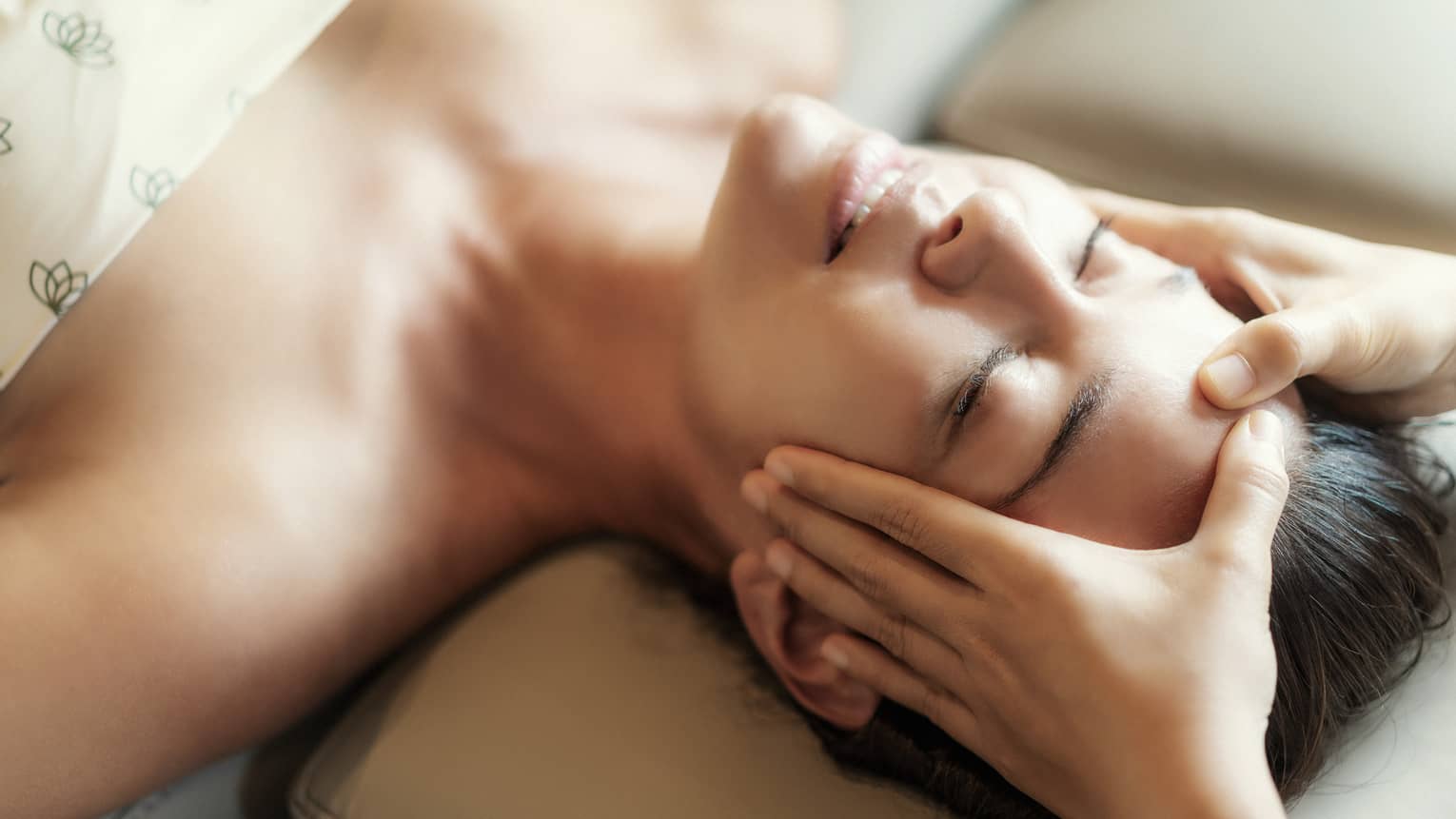 Spa facial, two hands rest on woman's forehead as she closes her eyes, lays under sheet on treatment table