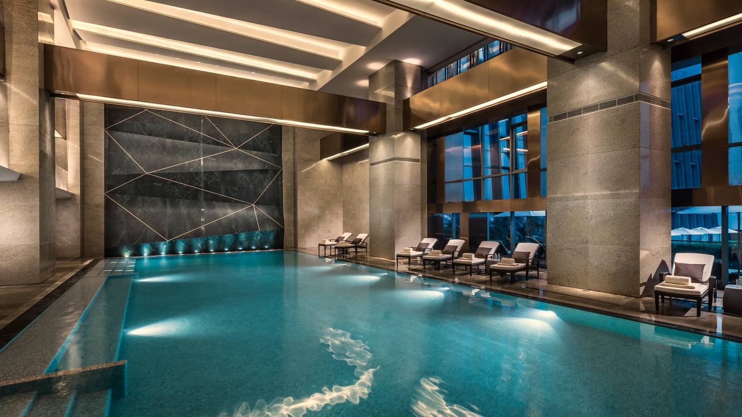 Lounge chairs line indoor swimming pool under marble pillars, ceiling lights