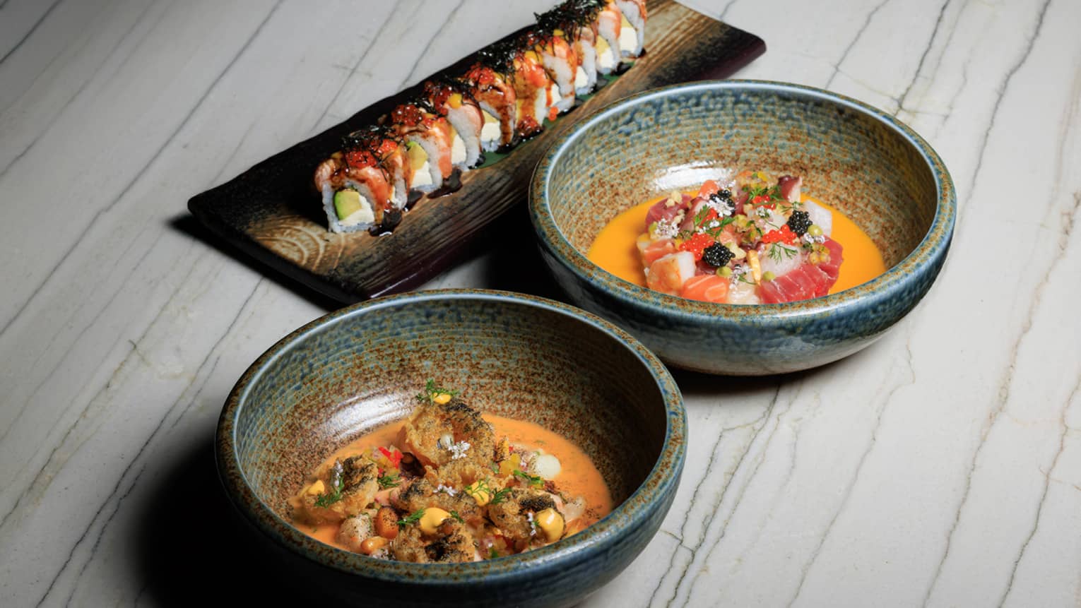 Two stoneware bowls filled with food sit next to a rectangular platter of sushi on a white wood-grain table