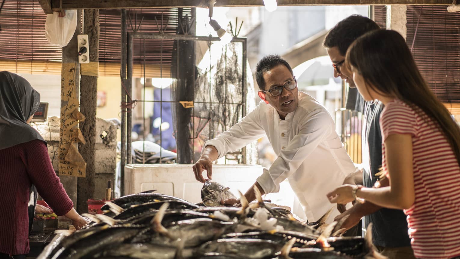 A merchant gently holds a fish with two hands while talking to two guests as they all stand by an indoor market fish table.