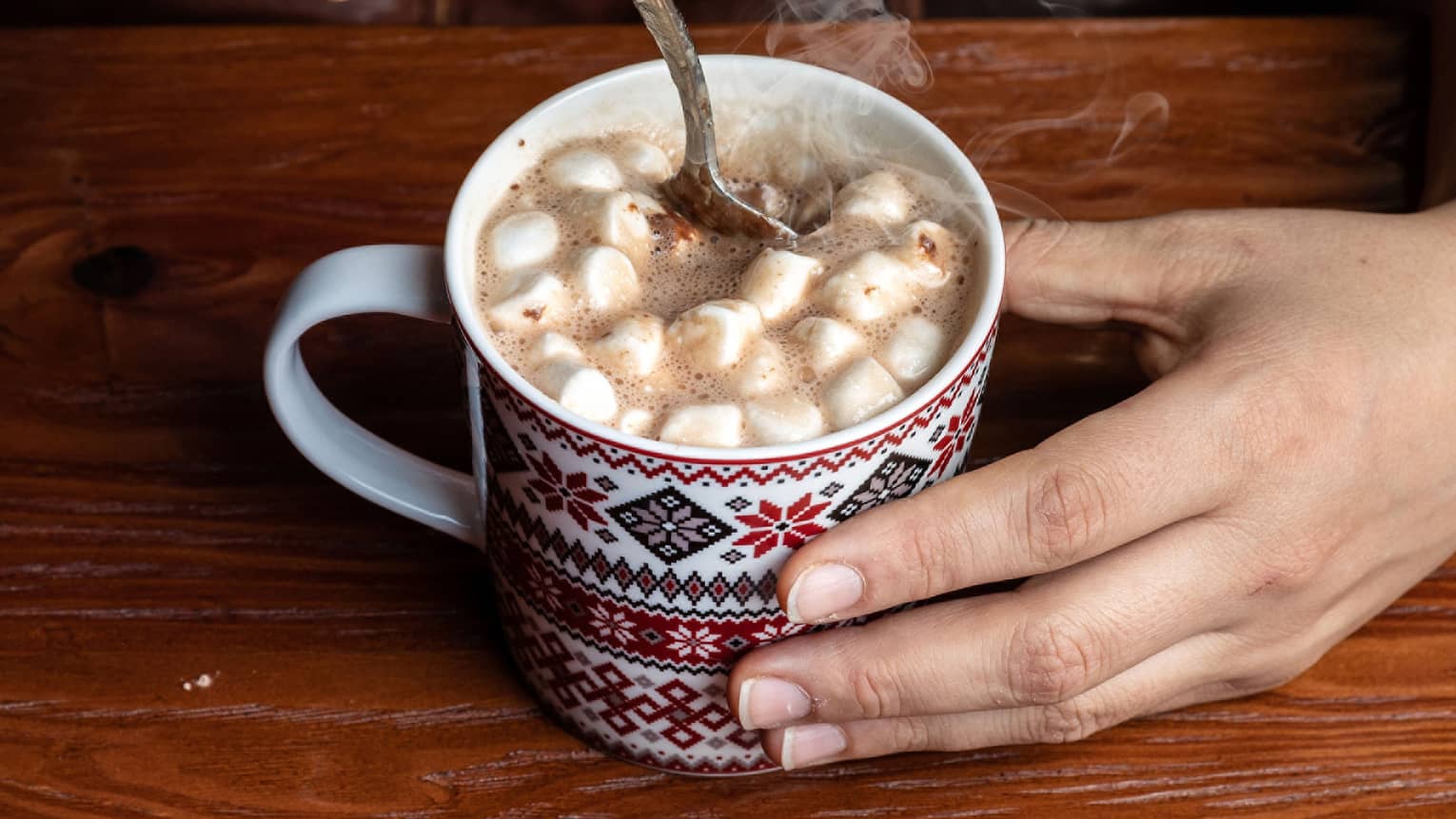Close up of two hands holding and stirring hot chocolate with marshmallows in a patterned mug