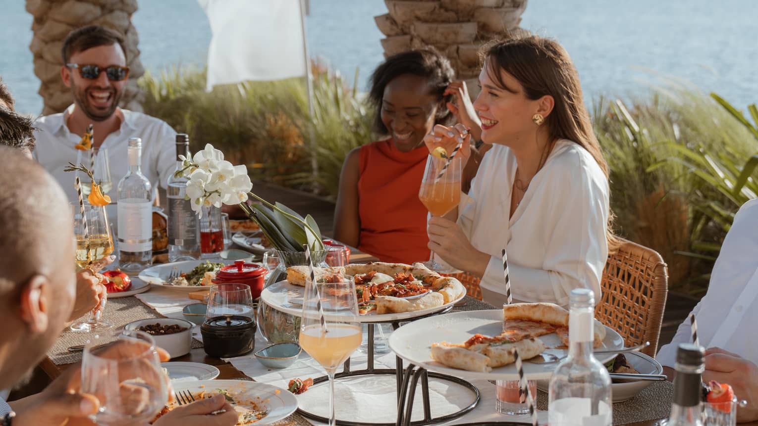 Group sits around long outdoor dining table eating brunch with the ocean and a city skyline in the background