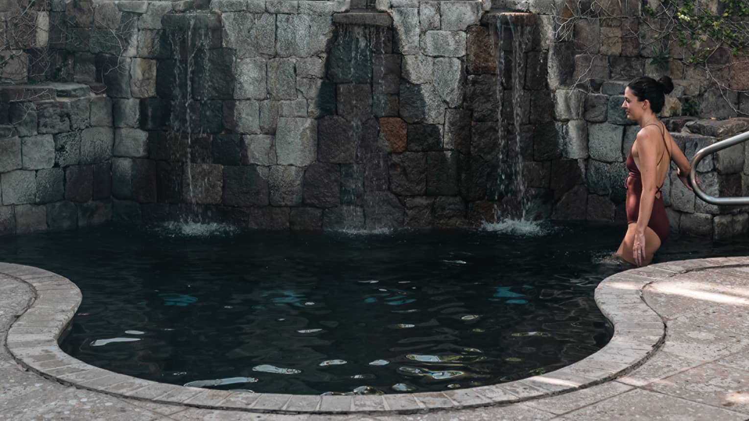 A woman getting into a small pool surrounded by stones and a small waterfall.