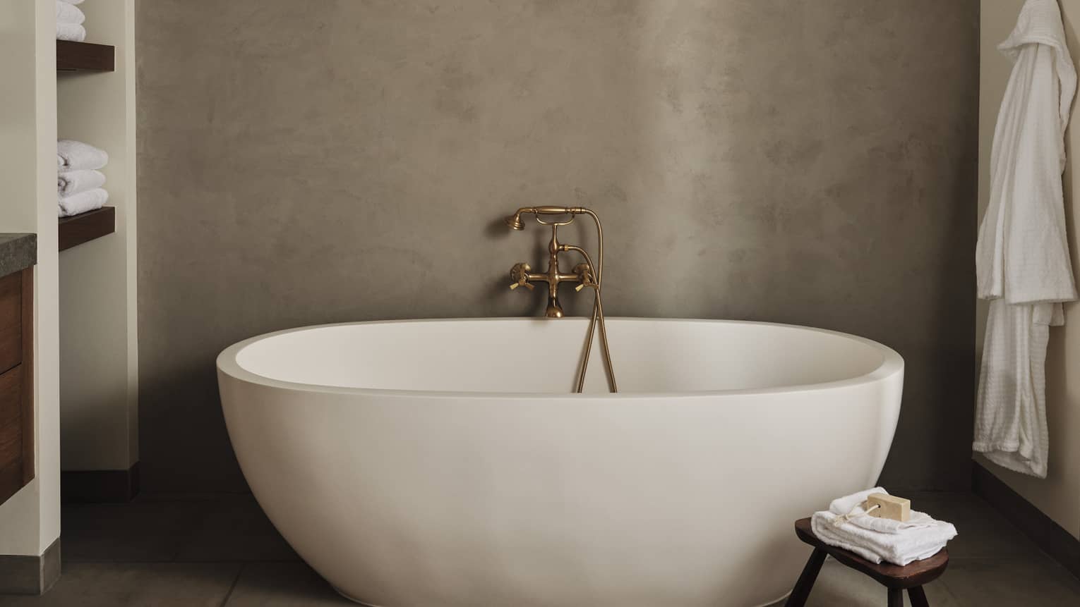 Luxury resort bathroom with a freestanding oval bathtub, built-in towel shelves, small wooden stool with folded towels and soap and bathrobe hanging on the wall