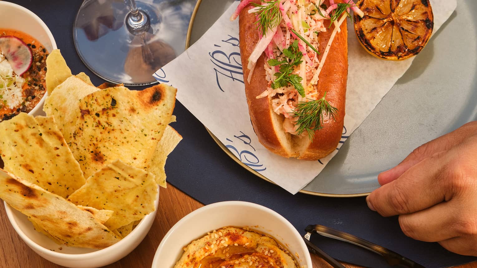 Hands reach for a hoagie-style sandwich served on a blue plate with various small bowls of dips and pita crips nearby