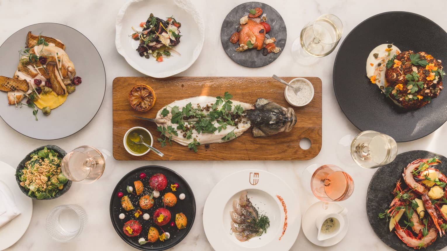 Aerial view of dining table with gourmet entrees, whole fish on wood platter, roasted seafood and vegetables