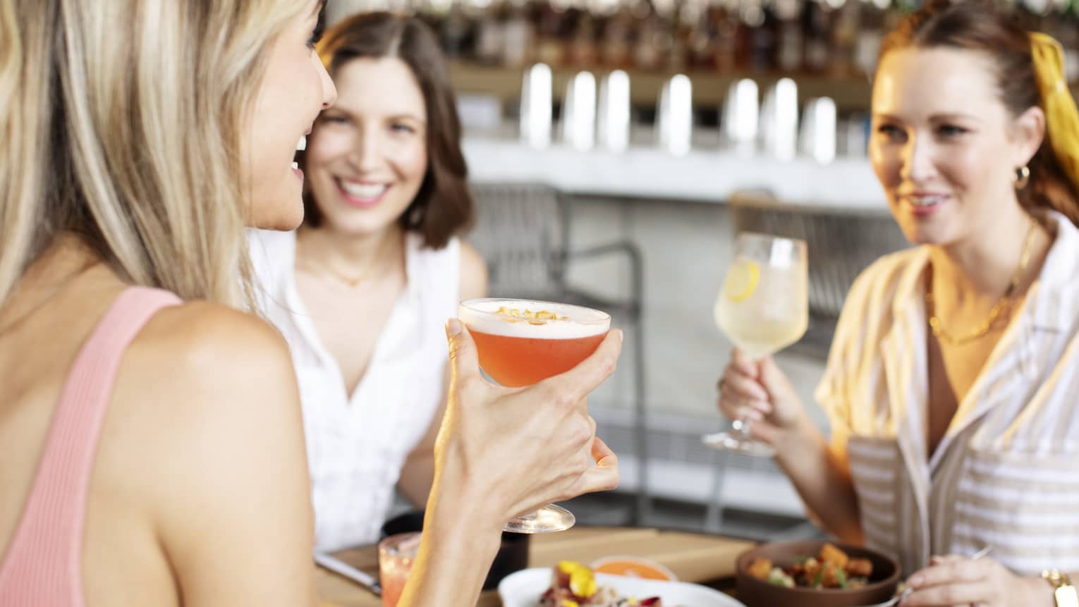 Three people wearing light, summery clothing sit around an outdoor table smiling and drinking cocktails