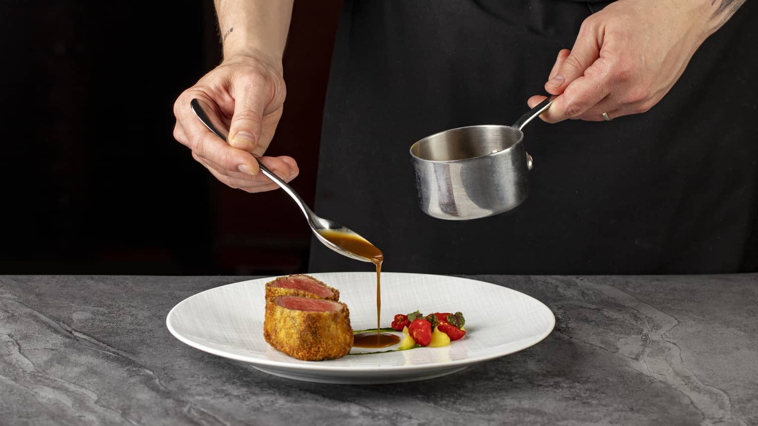 Chef?s hand spoons demi glace from metal measuring cup onto centre of white plate