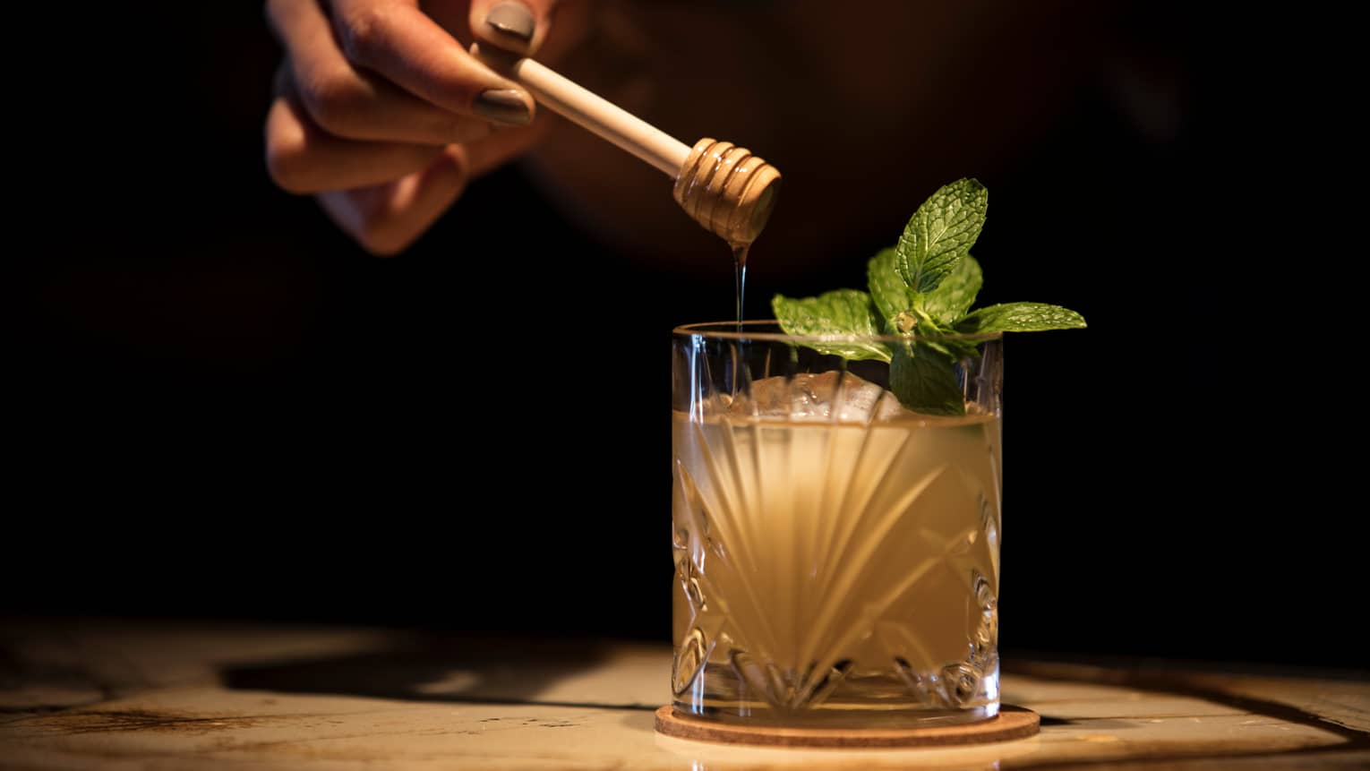 A cocktail with a sprig of mint sits on a table with dim lighting, a hand drizzles honey into it
