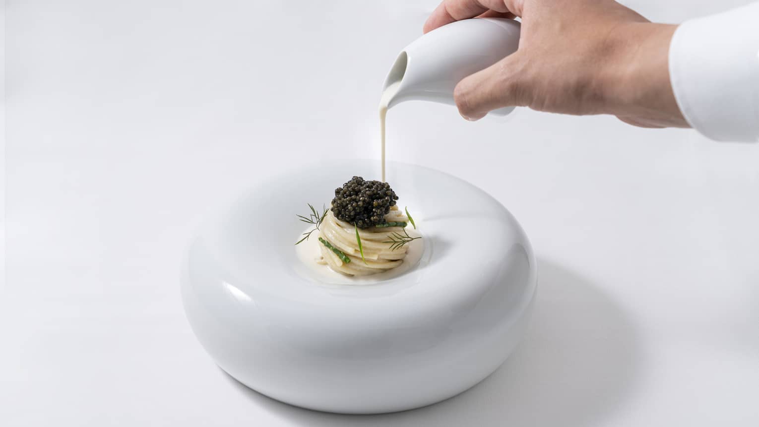 A chef's hand pours sauce onto "Caviar Pasta" set on modern white plate