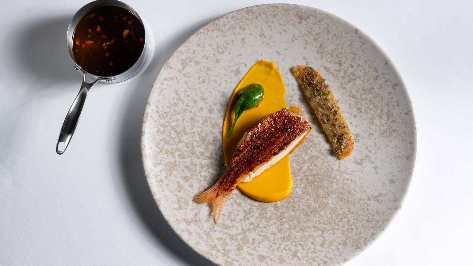 Red Mullet Bourguignon with Smoked Eel, Pearl Onion and Potato Galette on tan stone plate with measuring cup on side