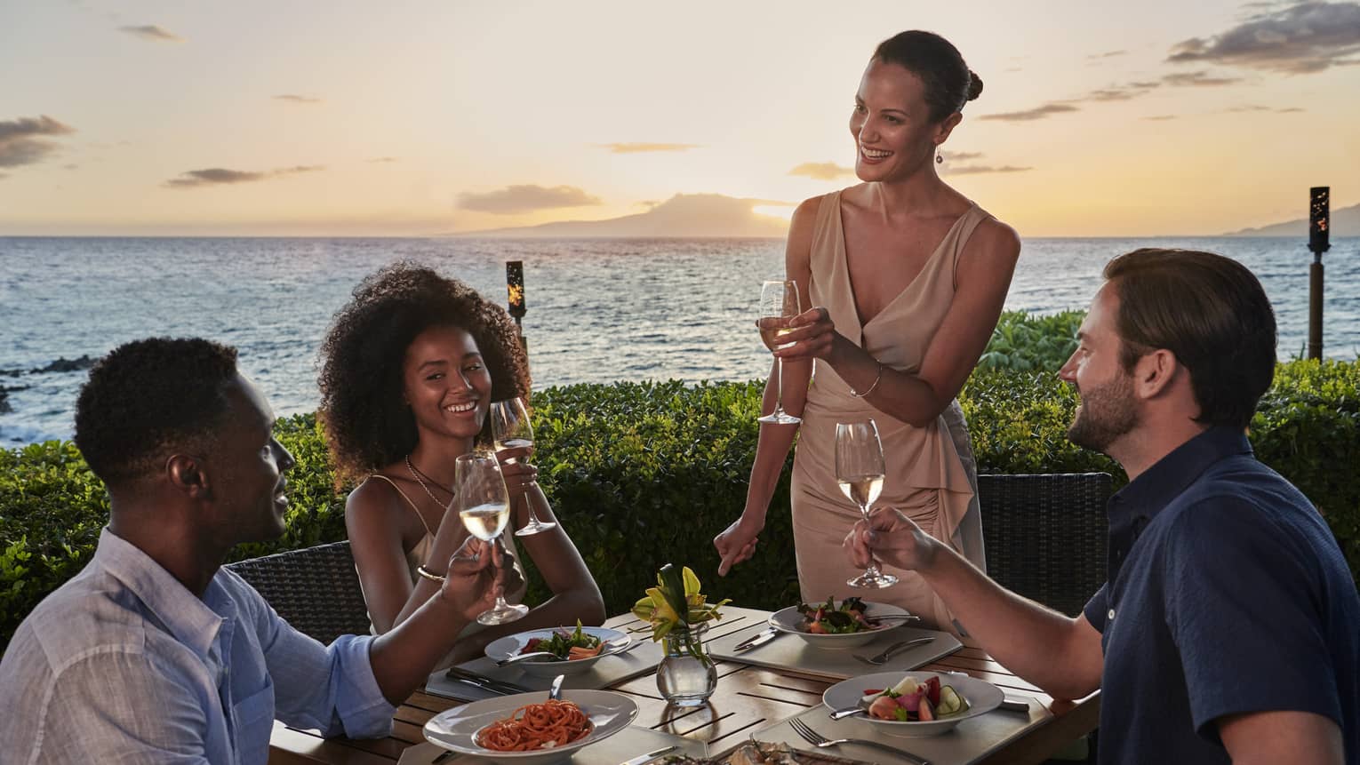 Two couples dine outside at Ferraro’s Bar E Ristorante, overlooking Wailea Beach. The sun is setting as they raise a toast. 