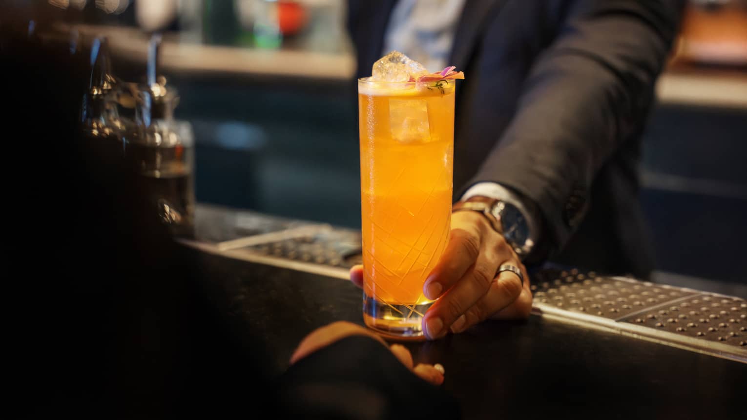 A person sets a bright orange drink in a tall clear glass on a bar counter.