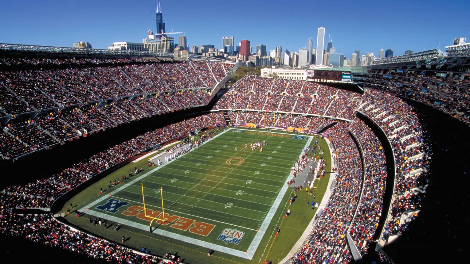 Aerial view of open stadium with football field, stands filled with fans against blue sky