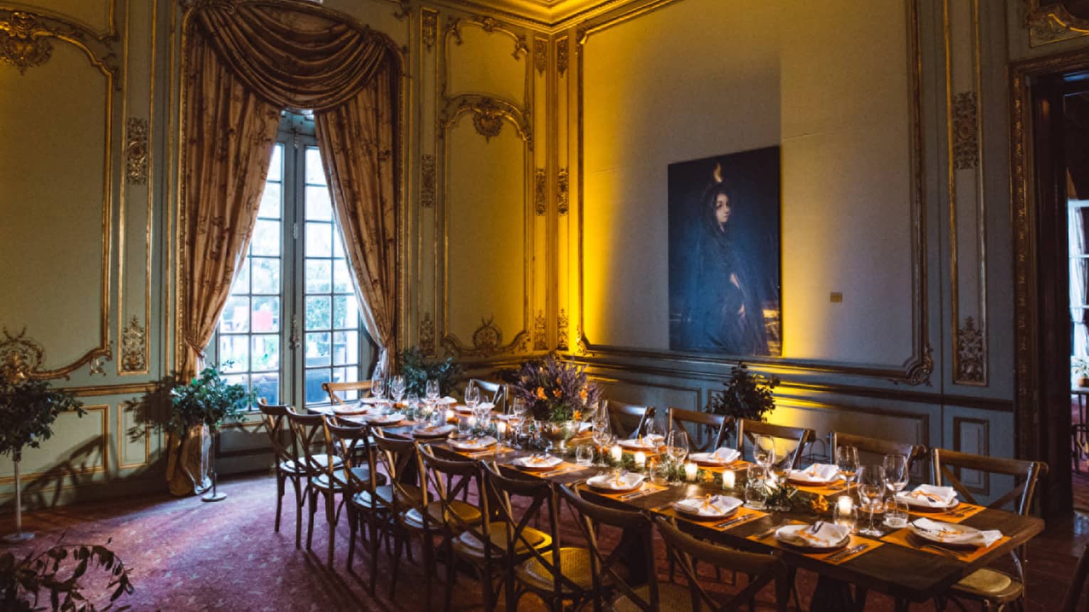 A long rectangular table is set for dinner in a dimly lit dining room with ornate drapes, oriental rugs and a large window
