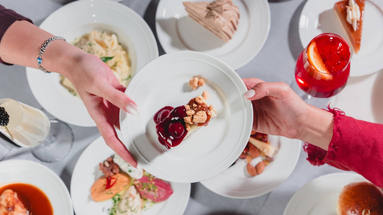 One hand passes along a white plate to another hand with a variety of dishes of food seen below on a table