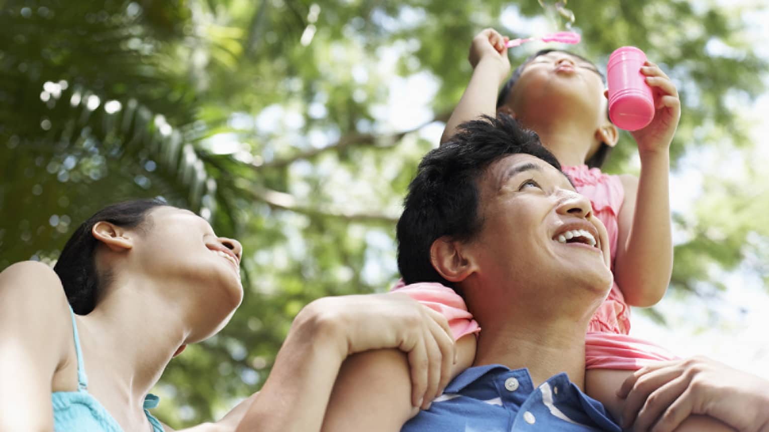 Young girl blows bubbles with pink wand as she sits on laughing man's shoulders, smiling woman beside him
