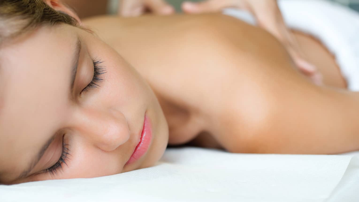 Woman with eyes closed rests on spa treatment table while massage therapist places hands on her bare back