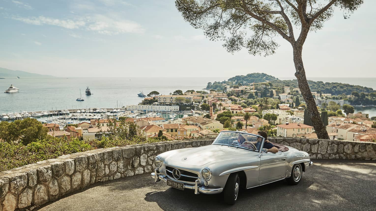 Couple in luxury vintage white car parked under tree by stone wall overlooking red roofs, water