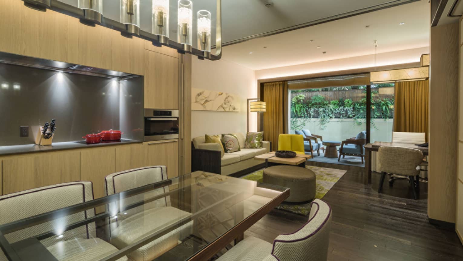 Luxury residential suite with dining table, kitchenette, sofa, desk and chairs, leading to a patio with outdoor seating and view of lush greenery