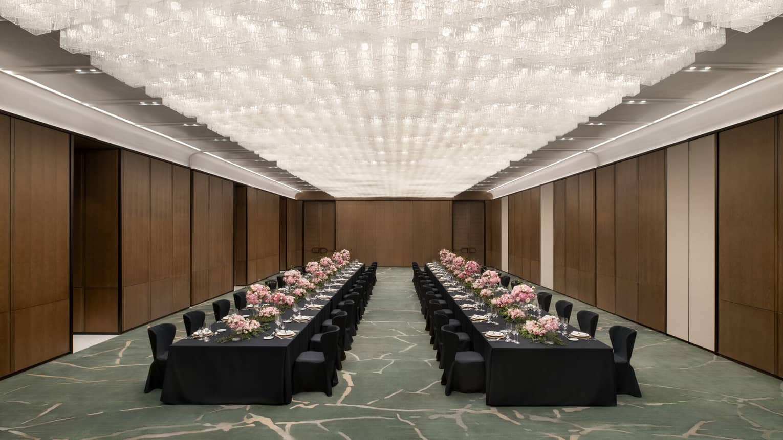 A spacious ballroom with two long black banquet tables set with plates, glassware, linens and pink floral arrangements
