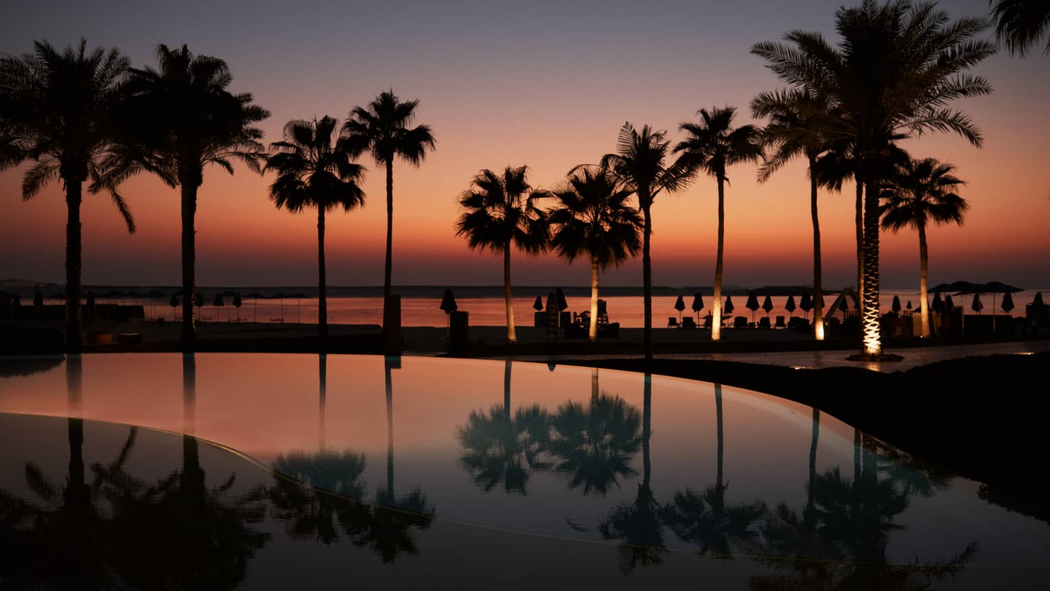 A palm tree?lined pool with the beach in the background all lit under a dark orange and blue sky as the sun sets