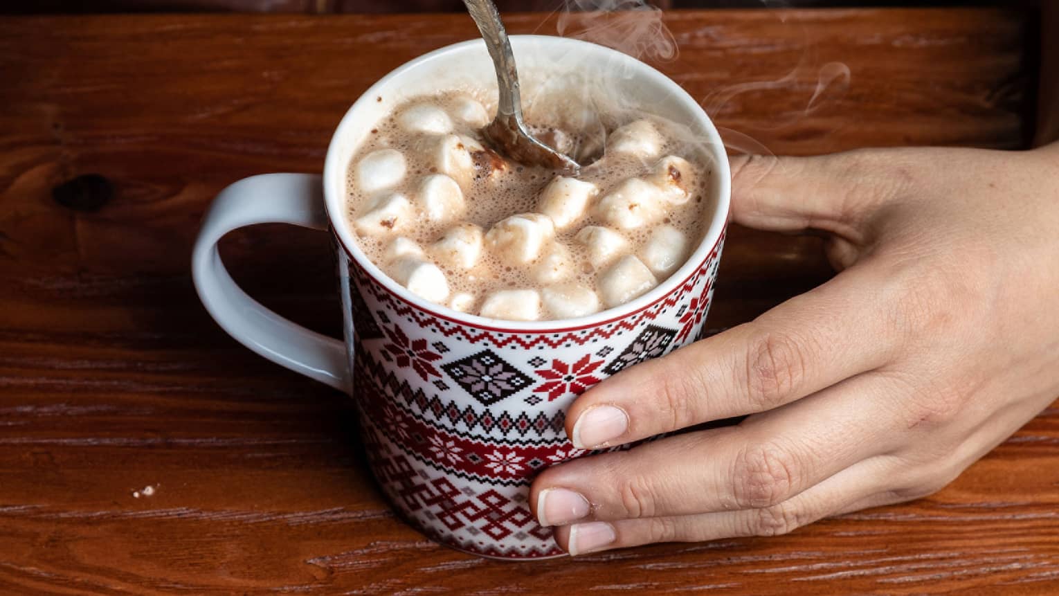 Close up of two hands holding and stirring hot chocolate with marshmallows in a patterned mug