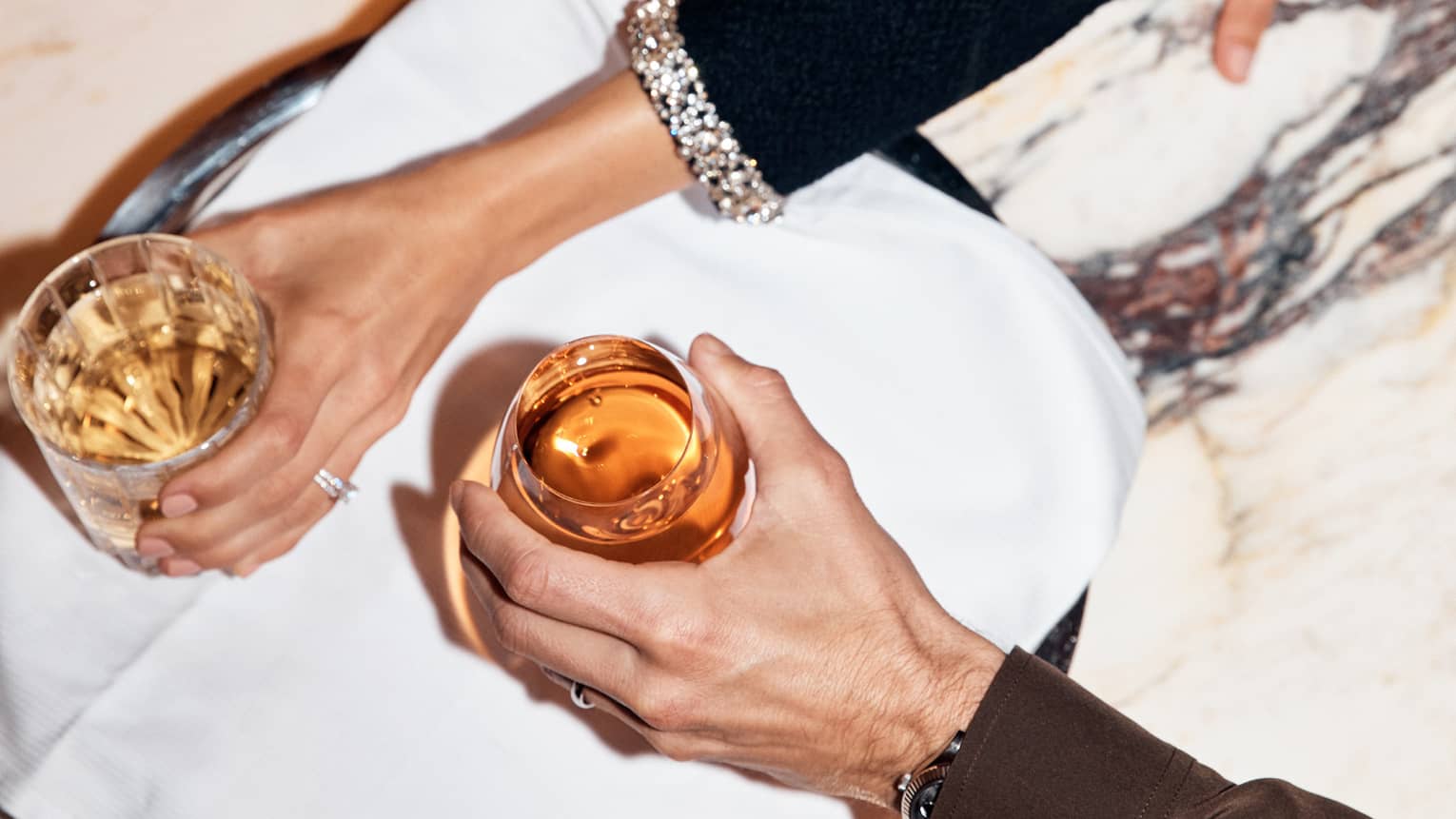 Two arms each reach out for a cocktail on a silver tray held above a white marble table