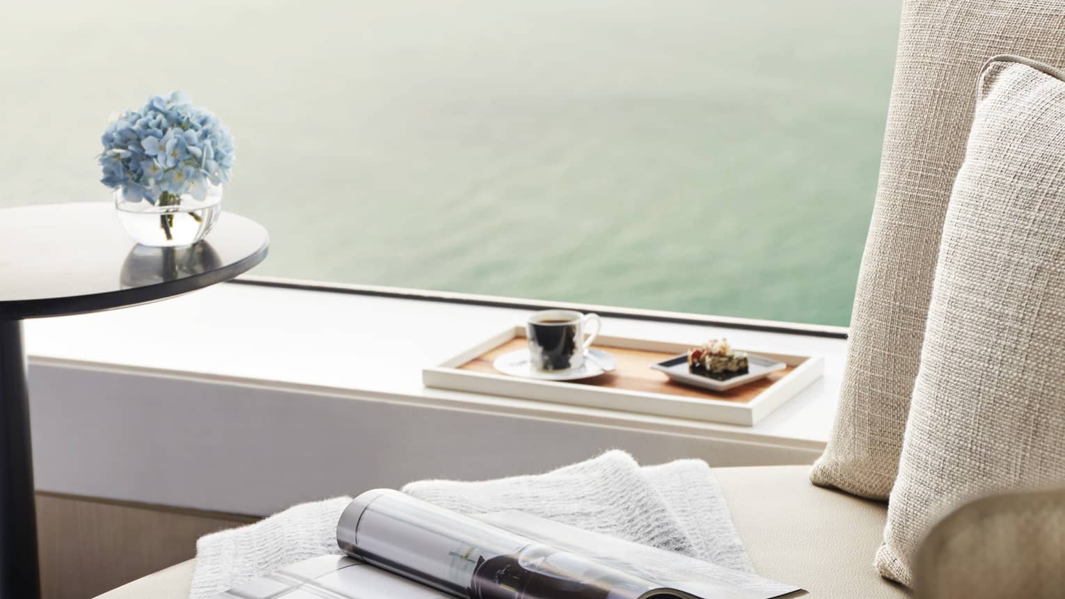 Close-up of cozy seating by window with a cup of coffee, pastries on a tray, a magazine, and blue flowers, overlooking water.