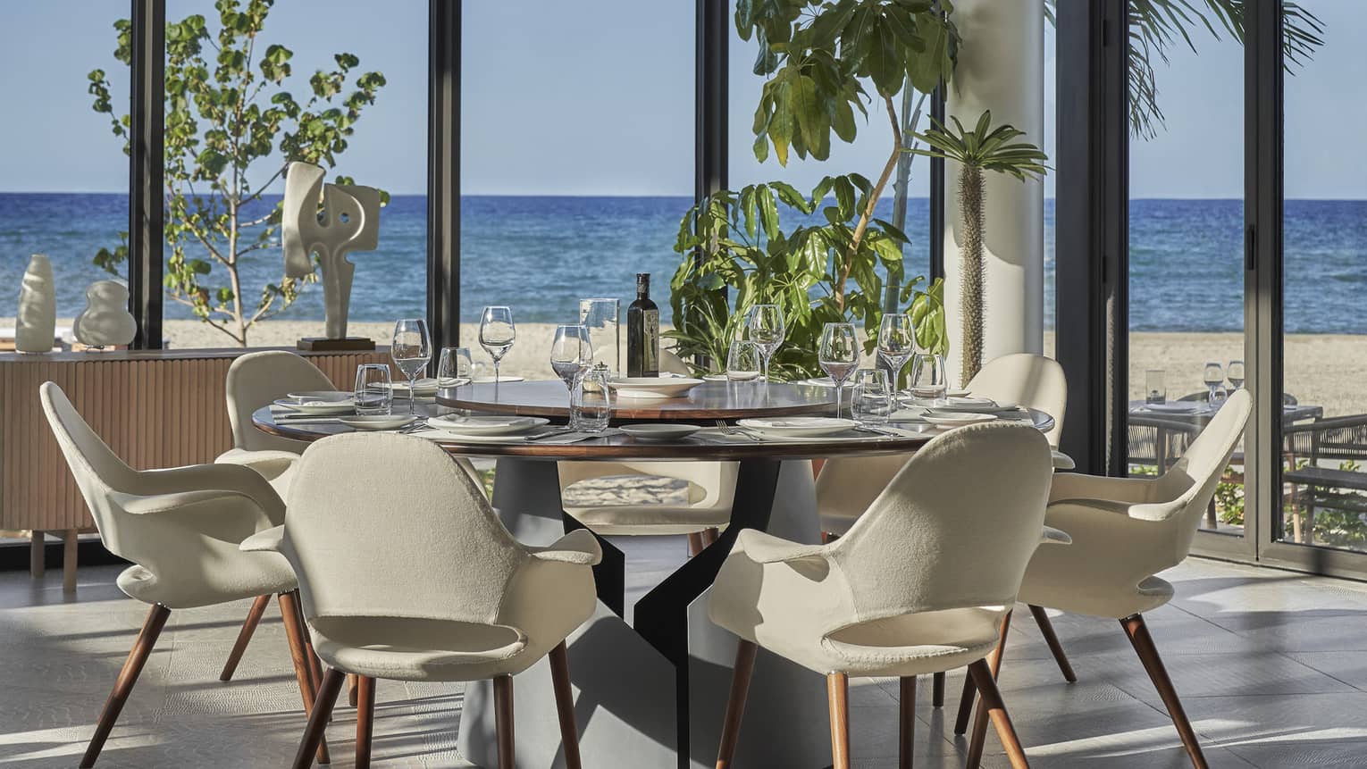 A round table with white leather chairs surrounding it and large floor to ceiling windows looking out at a beach.
