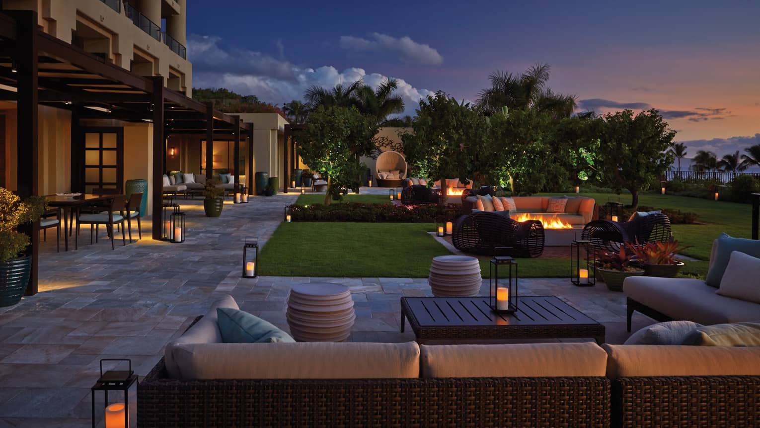 Outdoor lounge area at dusk with wicker seating, fire pits, lanterns and garden landscaping