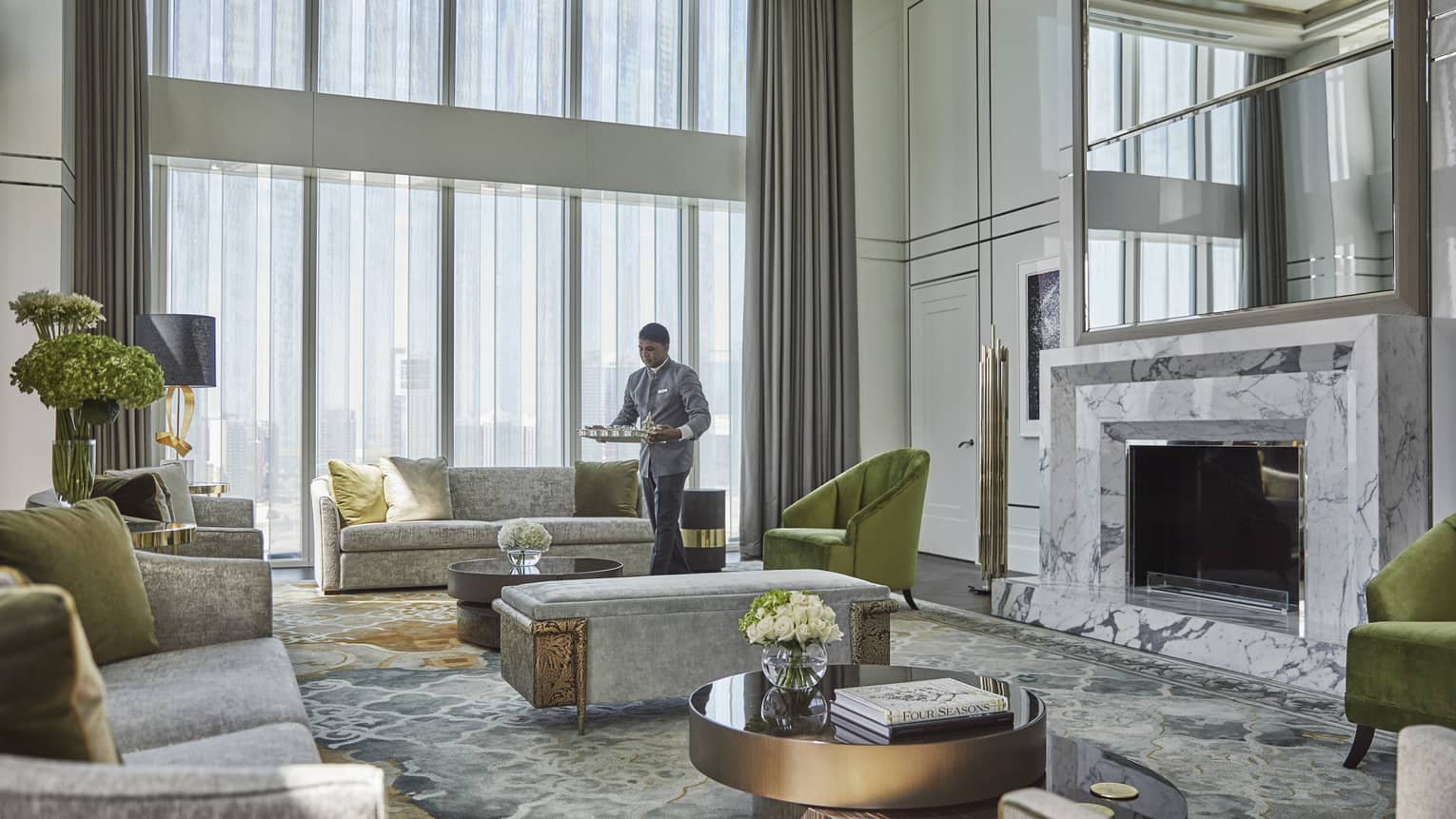Luxurious hotel living room with high ceilings, marble fireplace, plush gray sofas, green accent chairs, floral arrangements and an attendant serving refreshments