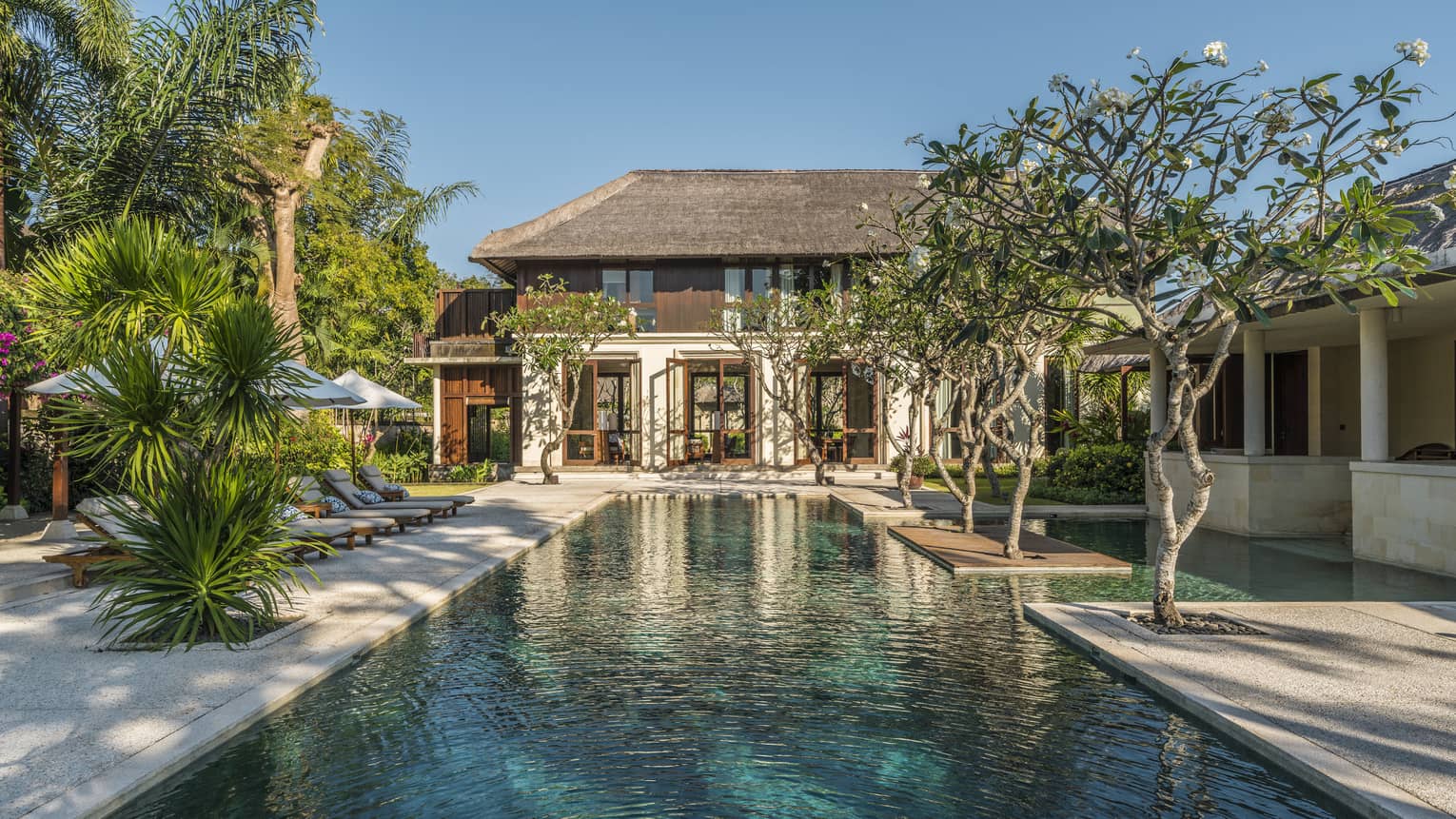 Two-storey villa at end of long outdoor swimming pool lined with trees, tropical plants